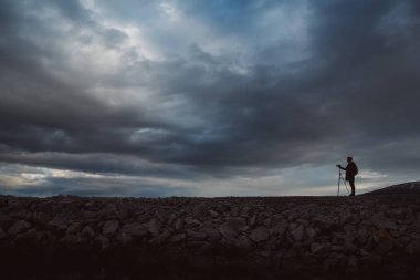 Bir fotoğrafçı veya gezgin siluet i tripod taş üzerinde duran. Dramatik bir gökyüzünün arka planı. Tehlikeli çalışma koşulları ve insan silueti