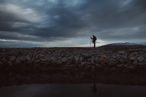 Bir fotoğrafçı veya gezgin siluet i tripod taş üzerinde duran. Dramatik bir gökyüzünün arka planı. Tehlikeli çalışma koşulları ve insan silueti