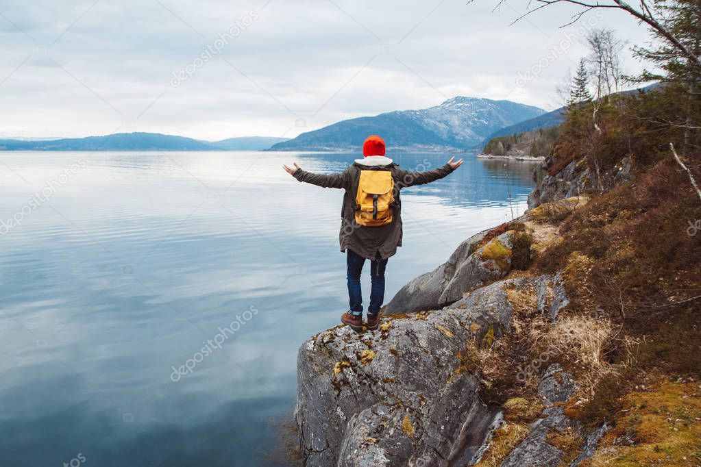 Traveler man with a yellow backpack wearing a red hat standing on a rock hands on the side on the background of mountain and lake. Space for your text message or promotional content. Travel lifestyle