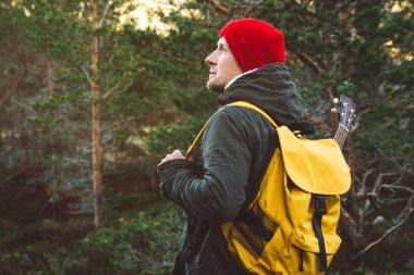 Gezgin adam bir gitar ile bir ormanın ortasında bir iz üzerinde duruyor. Kırmızı şapkalı sarı bir sırt çantası giyiyor. Lifestyle seyahat konsepti. Arkadan vur