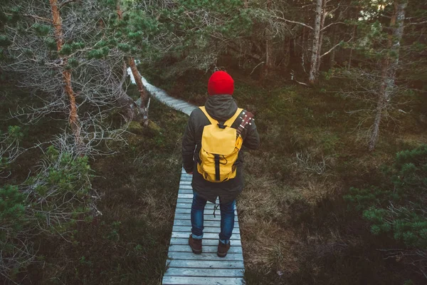 Traveler man is standing on a trail in the middle of a forest with a guitar. Wearing a yellow backpack in a red hat. Lifestyle Travel Concept. Shoot from the back — Stock Photo, Image
