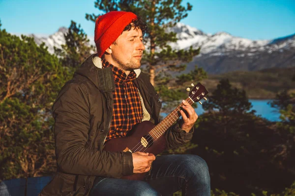 Guy playing guitar against the background of mountains, forests and lakes, wear a shirt and a red hat. Relaxing and enjoying sunny days.