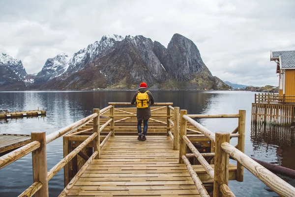 Ταξιδιώτης με ένα κίτρινο σακίδιο φορώντας ένα κόκκινο καπέλο στέκεται στο φόντο του βουνού και της λίμνης ξύλινη προβλήτα. ταξιδιωτικό τρόπο ζωής έννοια. Ρίξε από πίσω. — Φωτογραφία Αρχείου