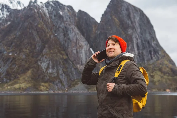 Portrait traveler man talking on mobile phone. Tourist in a yellow backpack standing on a background of a mountain and a lake. Traveler walks, smiling man calling friends. Telephone conversation.