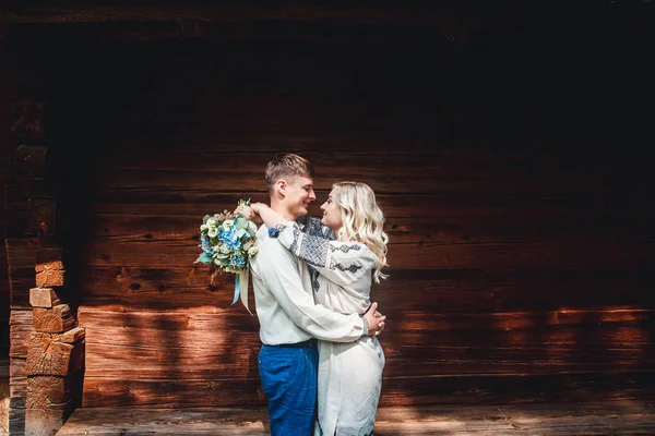 Couple de mariage incroyable dans une chemise brodée avec un bouquet de fleurs sur le fond d'une maison en bois — Photo