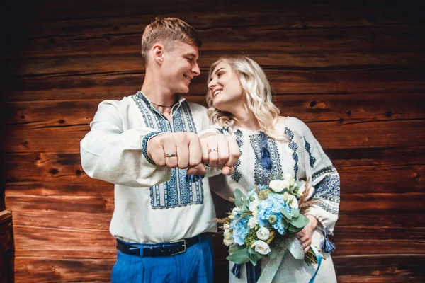 Amazing wedding couple in an embroidereds shirt with a bunch of flowers on the background of a wooden house — Stock Photo, Image