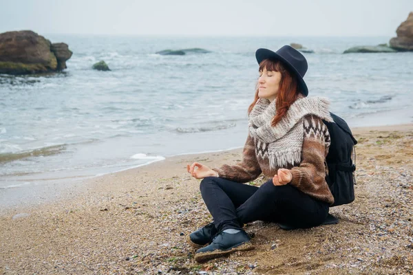 Bir şapka ve bir sırt çantası ile eşarp güzel kızıl saçlı kadın güzel denize karşı kayaların arka planına karşı sahilde meditative pozisyonda oturur. Turizm, dinlenme, yaşam tarzı.