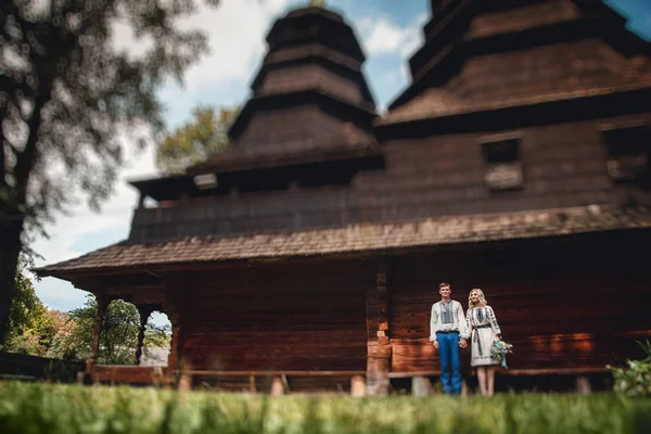 Amazing wedding couple in an embroidereds shirt with a bunch of flowers on the background of a wooden house — Stock Photo, Image