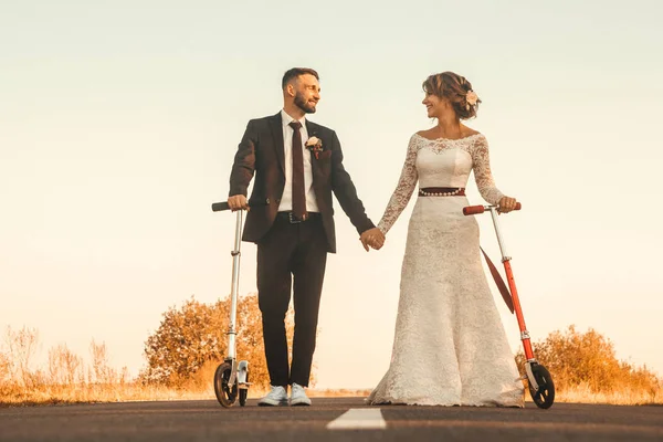 Couple de mariage souriant chevauchant un scooters sur la route en dehors de la ville au coucher du soleil. — Photo