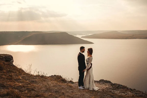 Incredibile matrimonio coppia, sposa e sposo che si tiene per mano su una montagna e fiumi sfondo. Bella ragazza in abito bianco, uomini in giacca e cravatta. Montagne paesaggio e fiumi al tramonto. Concetto di — Foto Stock