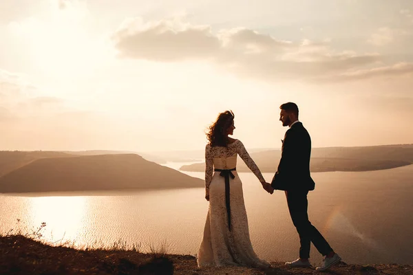 Increíble pareja de boda, novia y novio tomados de la mano en un fondo de montañas y ríos. Linda chica en vestido blanco, los hombres en traje de negocios. Montañas paisaje y ríos en la puesta del sol. Concepto de —  Fotos de Stock