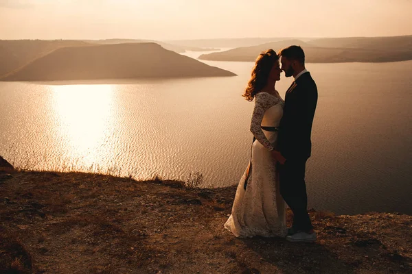 Increíble pareja de boda, novia y novio tomados de la mano en un fondo de montañas y ríos. Linda chica en vestido blanco, los hombres en traje de negocios. Montañas paisaje y ríos en la puesta del sol. Concepto de —  Fotos de Stock