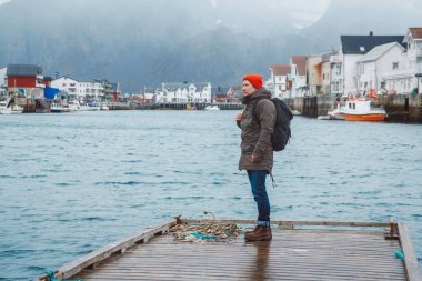 Sırt çantalı gezgin balıkçı kulübelerinin, gemilerin, dağların ve göl ahşap iskelede duran kırmızı bir şapka takıyor. Seyahat yaşam tarzı konsepti.