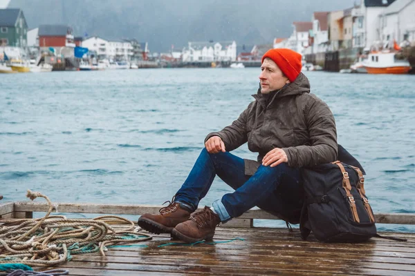 Kırmızı şapkalı bir sırt çantalı gezgin balıkçı kulübelerinin, gemilerin, dağların ve göl ahşap iskelelerinin arka planında oturuyor. Seyahat yaşam tarzı konsepti.