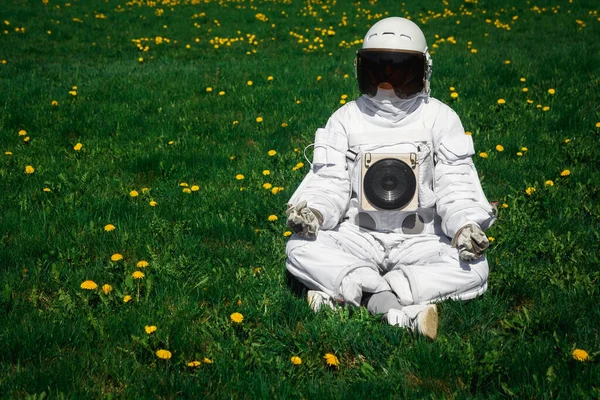 Futuristic astronaut in a helmet sits on a green lawn among flowersin a meditative position.