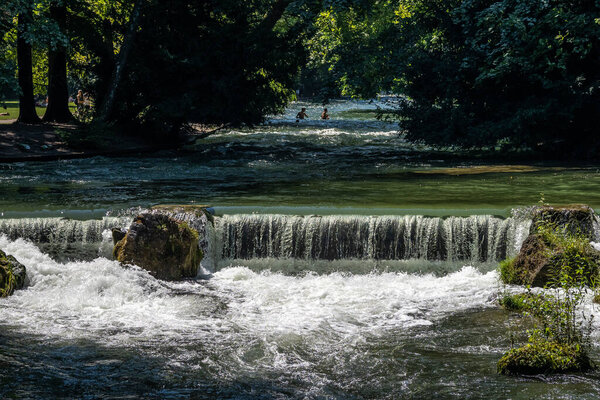water of the isar spilling over rocks of green moss and surrounded with tall green trees, in The English Garden, Munich, Germany.