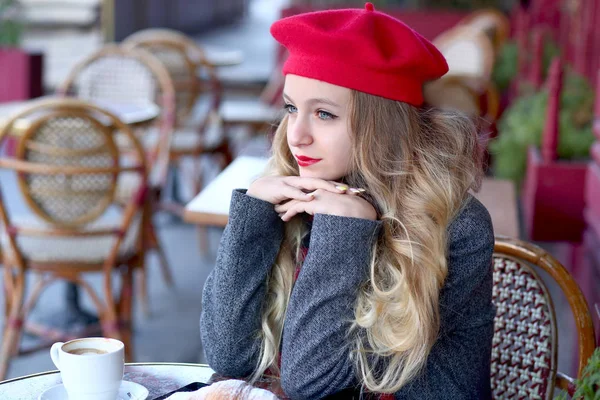 Jong en mals meisje in een rode baret zit aan een tafel en drinkt koffie in een straat café — Stockfoto