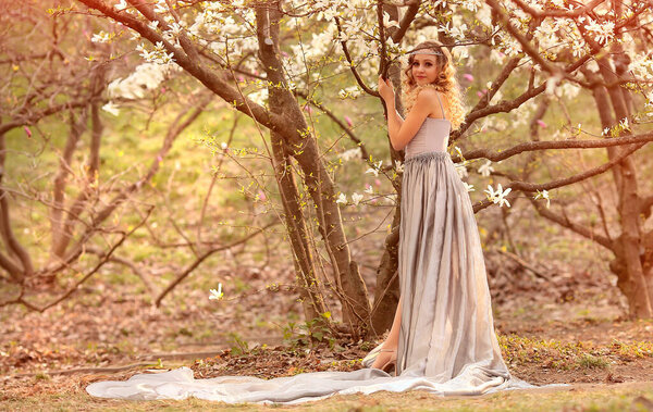 Young beauty blonde with jewelry for hair, in a silver dress with a long train in the garden where magnolia trees bloom. Beauty and fashion concept. Flares, bokeh effects.Copy space on the left