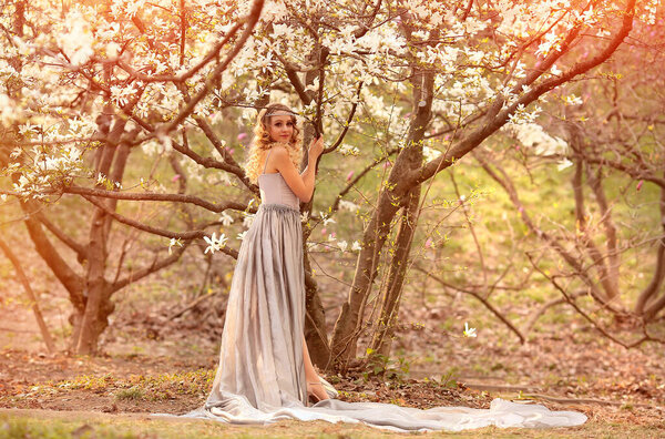 Young beauty blonde with jewelry for hair, in a silver dress with a long train in the garden where magnolia trees bloom. Beauty and fashion concept. Flares, bokeh effects.Copy space on the right