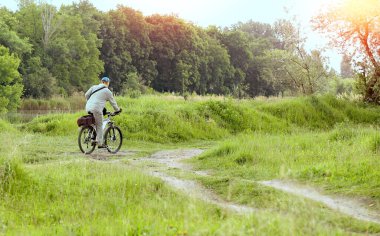 Gri eşofmanlı bir adam güzel turuncu bir gün batımında nehir kenarındaki bir ormanda bisiklet sürüyor. Doğa, ışınlar, boke. Etkin yaşam tarzı kavramı