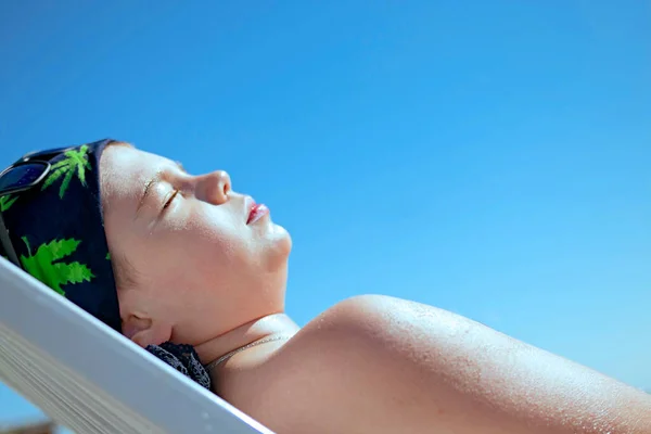 Teenager boy lying in the sun closing his eyes sunbathes on a sea beach. Caucasians attractive child on a blue sky background, child\'s face close-up. Cosmetics for the beach. Vacation concept