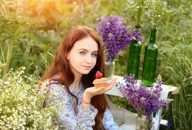 Genç bir kız doğada oturur ve mavi elbiseli kameraya bakar ve çilekli kek yer. Piknik ve açık hava eğlencesi. Yaz konsepti.