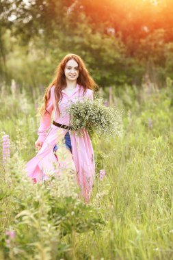 Büyük bir buket papatyayla pembe elbiseli güzel kızıl saçlı kız yazın gün batımında bahçede koşar. Bokeh, Flash, güneş ışınlarının etkisi. Yaz kavramı. Boşluğu kopyala