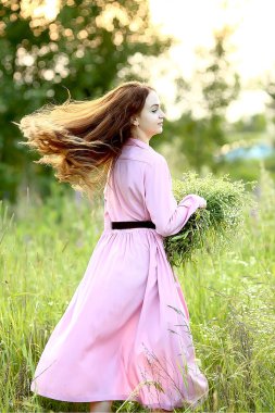 Pembe elbiseli güzel kızıl saçlı kız gün batımında bahçede kocaman bir buket yaz papatyası ile dönüyor. Bokeh, Flash, güneş ışınlarının etkisi. Yaz. Rahatlama konsepti