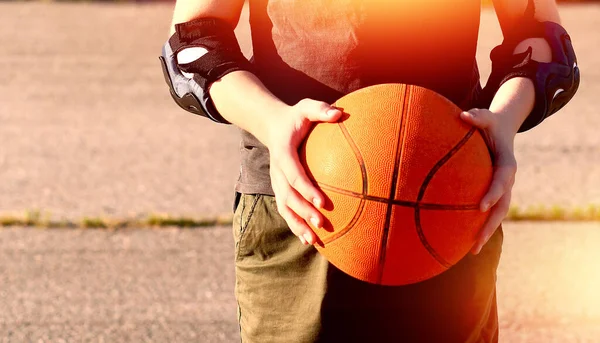 Orange basketball ball in the hands of a teenager on the background of the sports ground on a summer day. Close-up. Copy space on the left