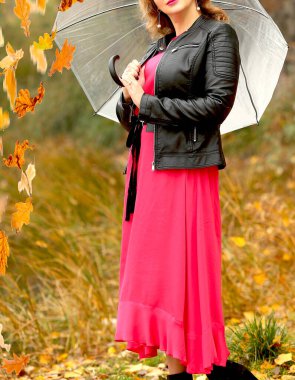 Siyah deri ceketli, kırmızı şık elbiseli ve şemsiyeli genç bir kız parkta yürüyor. Ağaçlardan yapraklar dökülüyor. Sonbahar. 