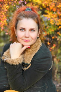 Genç bir kız, sonbahar parkında kırmızı kuyruklu ve sakin bir şekilde kameraya yakından bakıyor. Kız sarı kot pantolon ve yeşil ceket giyiyor. Sonbahar kadın kıyafetleri. Sonbahar konsepti