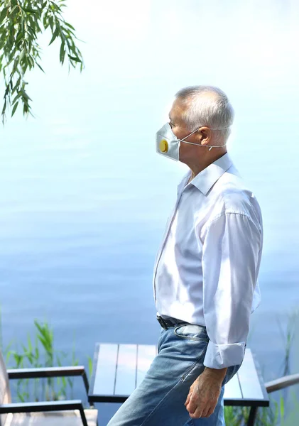 A mature man in protective respirator walks at a lake. A man over 60 years old with gray hair in a white shirt. Elderly lifestyle. Coronavirus protection rules.