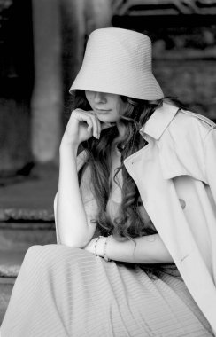 Siyah beyaz fotoğraflı, bej renkli trençkotlu, mavi elbiseli, kova şapkalı, spor ayakkabılı, geometrik desenli tahta kapının yanında poz veren genç bir kadın. Şehir tarzı sokak portresi