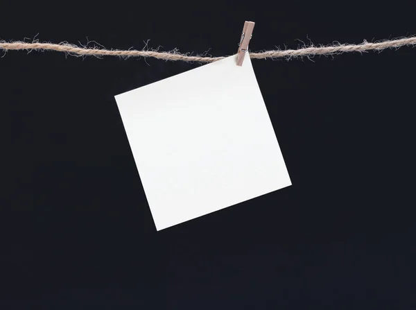 A white square piece of note paper hangs on clothespins on a black background, mock up — ストック写真