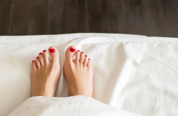 Dedo femenino con pedicura roja en la cama de debajo de las cubiertas blancas Primer plano de los pies femeninos cubiertos con pedicura roja — Foto de Stock