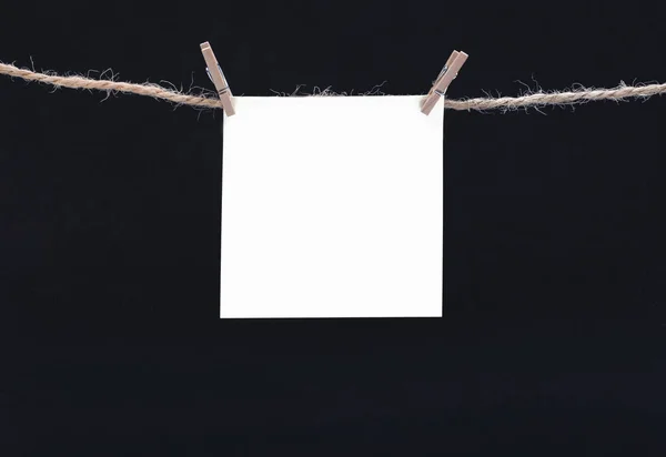 A white square piece of note paper hangs on clothespins on a black background, mock up — Zdjęcie stockowe