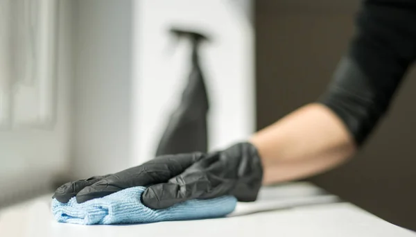 Uma mão em luvas pretas segura um pano de limpeza de microfibra e um spray com esterilização fazem a desinfecção para uma boa higiene — Fotografia de Stock