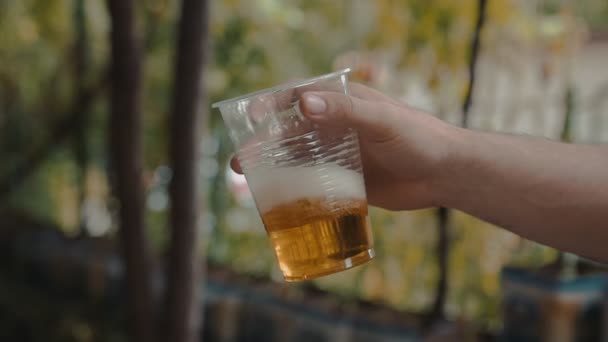 Мужская рука держит и медленно пожимает прозрачную пластиковую чашку со светлым пивом — стоковое видео