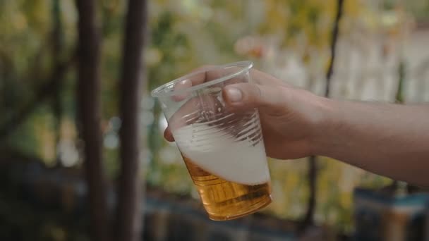 La mano del hombre sostiene y sacude lentamente una taza de plástico transparente con cerveza ligera — Vídeos de Stock