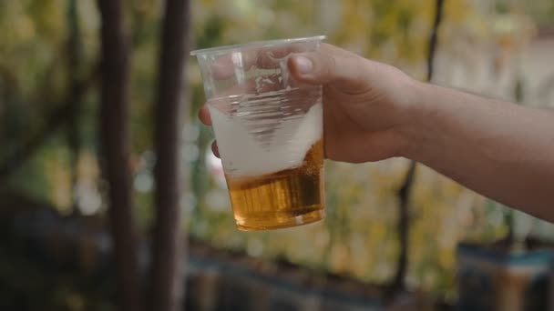 La mano del hombre sostiene y sacude lentamente una taza de plástico transparente con cerveza ligera — Vídeos de Stock