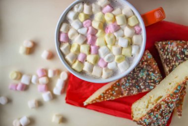 Cocoa drink with colorful small marshmallows and pieces of sweet homemade cake in a wooden tray.