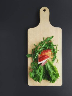 Kesme tahtasında taze yeşil marul ve roka yapraklarıyla dilimlenmiş jambon..