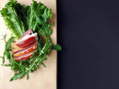 Kesme tahtasında taze yeşil marul ve roka yapraklarıyla dilimlenmiş jambon..