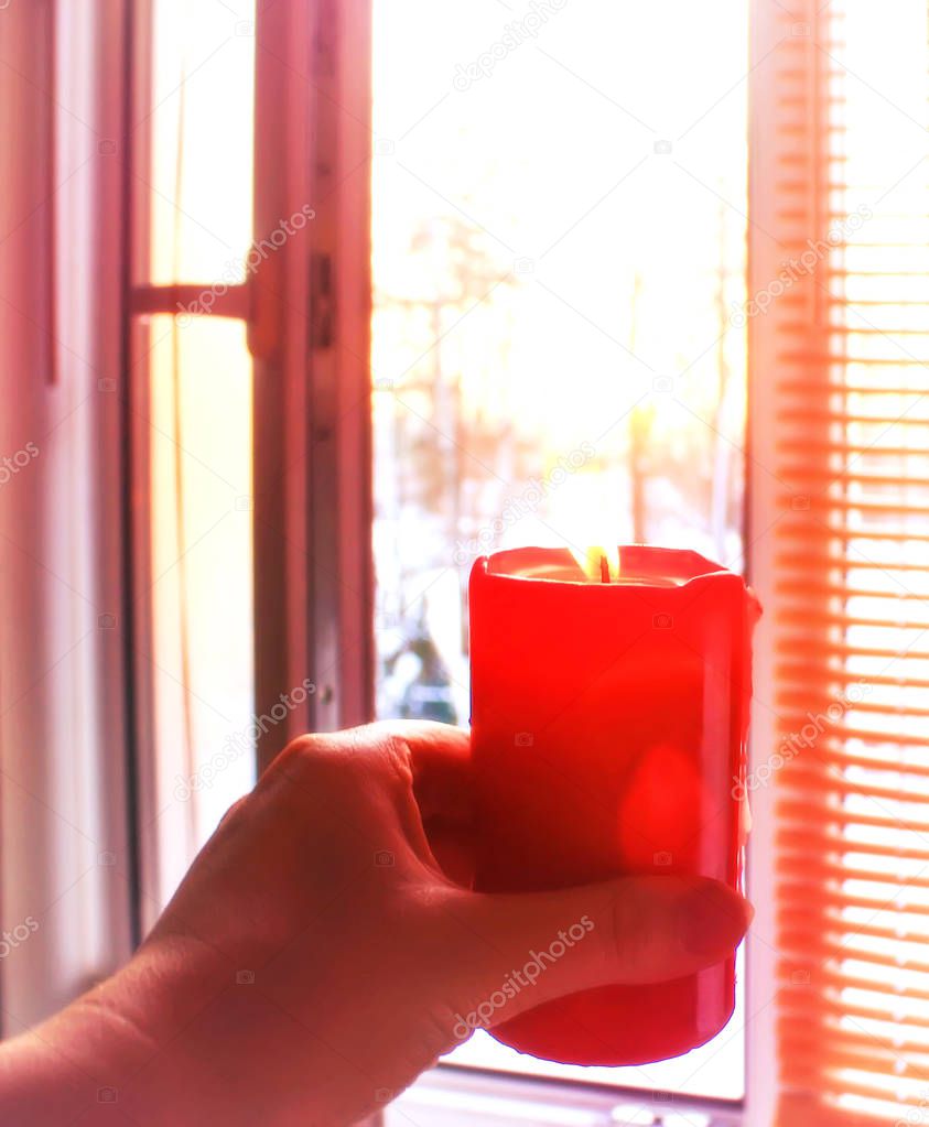 Red burning candle in a hand on window background.