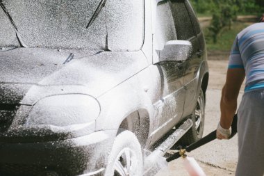Bir adam, arabaya yüksek basınç uygulayan bir temizlik maddesini püskürtüyor.