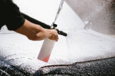 Bir adam, arabaya yüksek basınç uygulayan bir temizlik maddesini püskürtüyor.