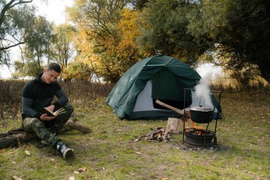 Ormandaki çadırın yanında bir adam deftere yazıyor.