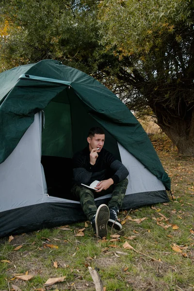 一个人在森林里的一个帐篷边的笔记本上写字 — 图库照片