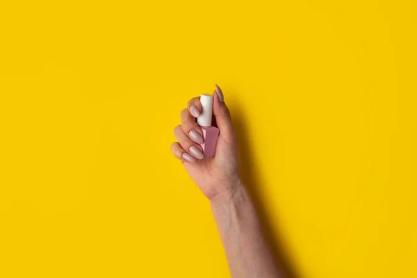 Gel polish of pink color in a hand on a yellow background, top view. Woman holds in hands pink bottle of varnish on a yellow background. Manicure concept