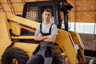 Tulum giymiş ve traktör arka planında kameraya gülümseyen genç bir çiftçinin portresi. Erkek traktör sürücüsü. Tarım, tarım ve ekoloji kavramı.